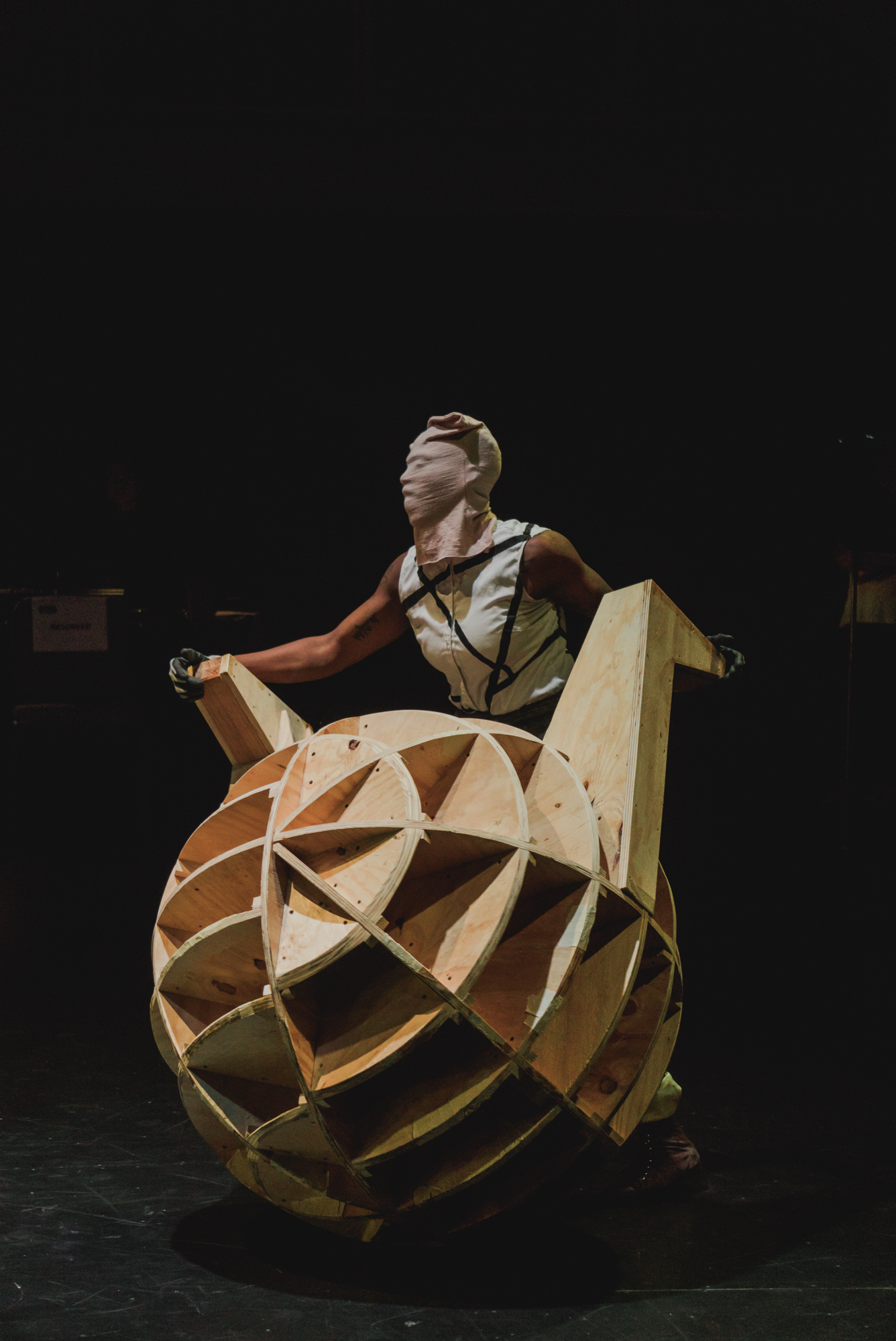 A performer with the Commons Choir stands masked behind a large spherical wooden construction